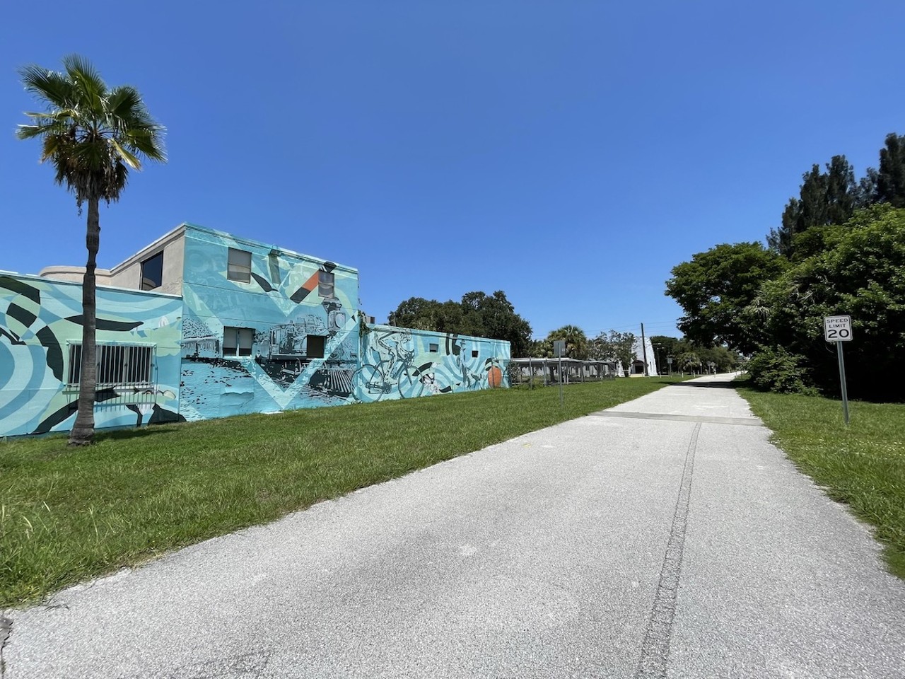 100 Years Before J. Cole tells the history of the Pinellas Trail in Downtown Clearwater
620 Drew St., Clearwater
You’ll find Tony Krol and Michelle Sawyer’s “100 Years Before J. Cole” alongside the Pinellas Trail, near where Drew Street intersects with North Garden Avenue.
The mural tells the history of the Pinellas Trail, once Peter Demen’s short-lived Orange Belt Railway. The Orange Belt provided a path for trains delivering Florida citrus, vegetables and passengers across central Florida before the Great Freeze in 1894-95. It became part of the Plant System in 1895, then Atlantic Coast Line Railroad in 1902. The train delivered passengers up and down the east coast before Amtrack took over in 1971 and discontinued passenger service to Pinellas County in the early 1980s.
Bert Valery and then-County Administrator Fred Marquis launched plans to convert the extinct railway to a bike trail after a car struck and killed Valery’s son while riding his bike down Belleair Causeway in 1983. The first stretch of trail opened in 1990.
Nearly 30 years later, in 2018, Krol and Sawyer told the tale in paint. View “100 Years Before J. Cole” through the ARTours Clearwater app and watch that history come to life.Photo by Jennifer Ring