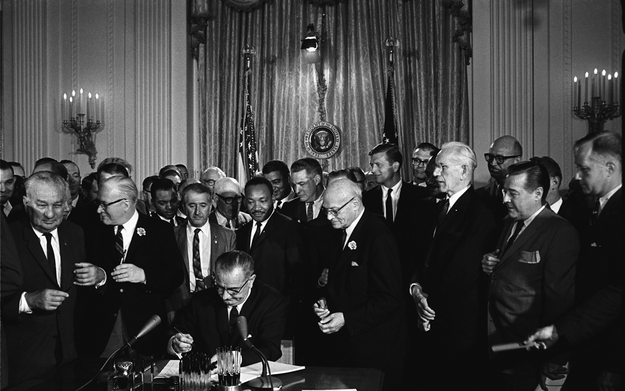 President Lyndon B. Johnson signs the Civil Rights Act of 1964. Martin Luther King, Jr.is among the many who are standing behind Johnson.