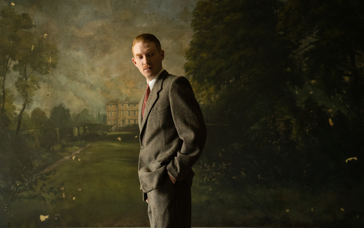 Dr. Faraday (Domhnall Gleeson) stands in front of a mural of Hundreds Hall, the possibly haunted mansion at the heart of The Little Stranger.